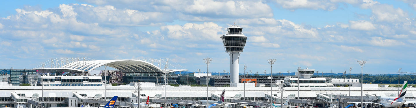 Arbeitgeber Flughafen Panoramablick auf den Flughafen München mit Terminalgebäuden, dem markanten Tower und mehreren abgestellten Flugzeugen unter einem hellen, leicht bewölkten Himmel; klare und ruhige Flughafenatmosphäre.