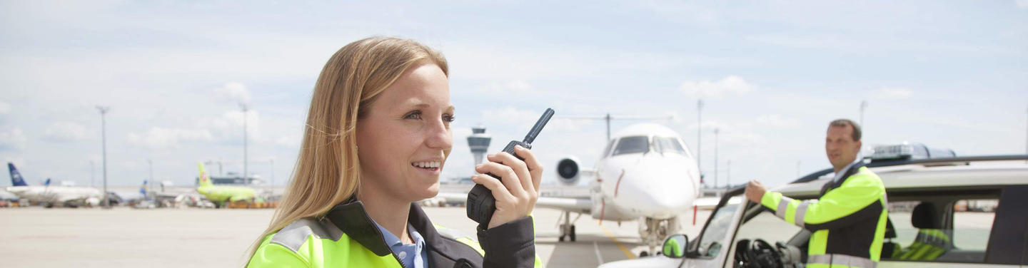 Schutz und Sicherheit am Flughafen München Eine Sicherheitsmitarbeiterin in Warnjacke spricht per Funkgerät auf dem Vorfeld des Flughafens München, im Hintergrund stehen ein Flugzeug und ein weiteres Einsatzfahrzeug; wachsame, professionelle und sicherheitsorientierte Atmosphäre.