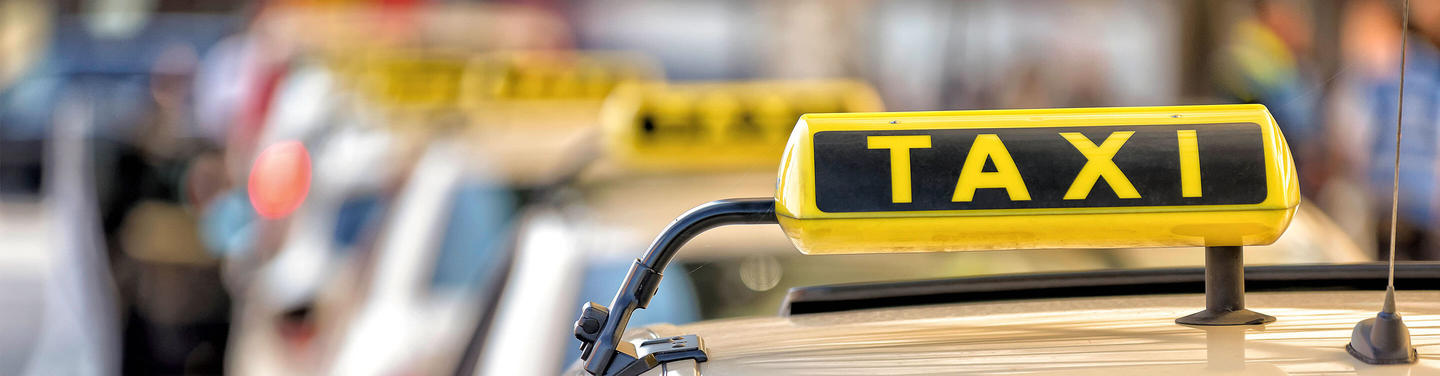 Taxi Service at Munich Airport Close-up of the yellow taxi roof sign with black “TAXI” lettering on a vehicle at Munich Airport; additional vehicles and people are visible out of focus in the background, creating a busy yet orderly atmosphere.