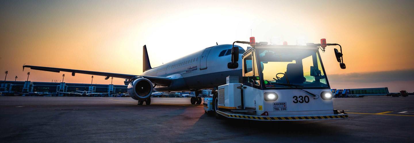 Flugzeugschlepper zieht Flugzeug Ein Flugzeug wird am Gate von Bodenfahrzeugen versorgt, während die Sonne in kräftigen Orange- und Blautönen hinter dem Flughafen untergeht. Die Szene wirkt ruhig, atmosphärisch und professionell.