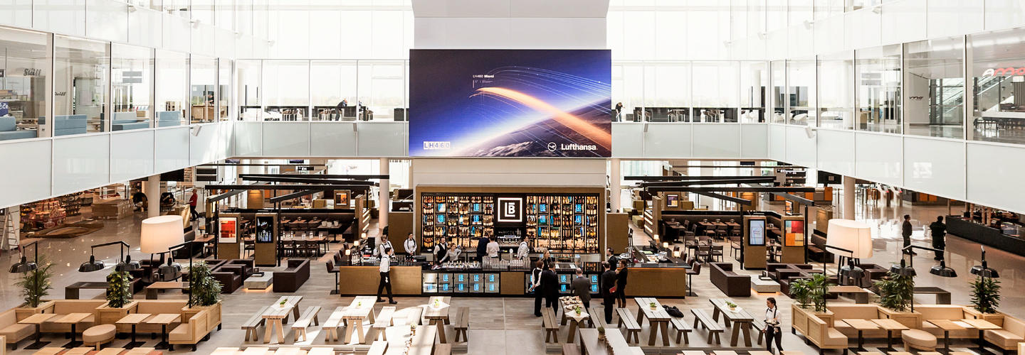 Werben bei Europas Nr. 1 Großer, offener Gastronomie- und Shoppingbereich im Terminal des Flughafens München mit modernen Sitzbereichen, Restaurants und einem großen LED-Screen. Die Szene vermittelt Vielfalt, Komfort und entspanntes Aufenthaltserlebnis für Reisende.
