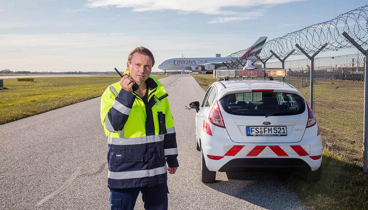 FMSicherheit Mitarbeiter mit Funkgerät am Flughafenzaun FMSicherheit Mitarbeiter mit Funkgerät am Flughafenzaun