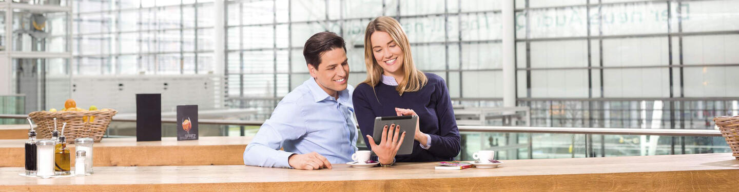 Couple with tablet Two people are sitting in a café area at Munich Airport, looking at a tablet together, with the glass facades of the terminal in the background.