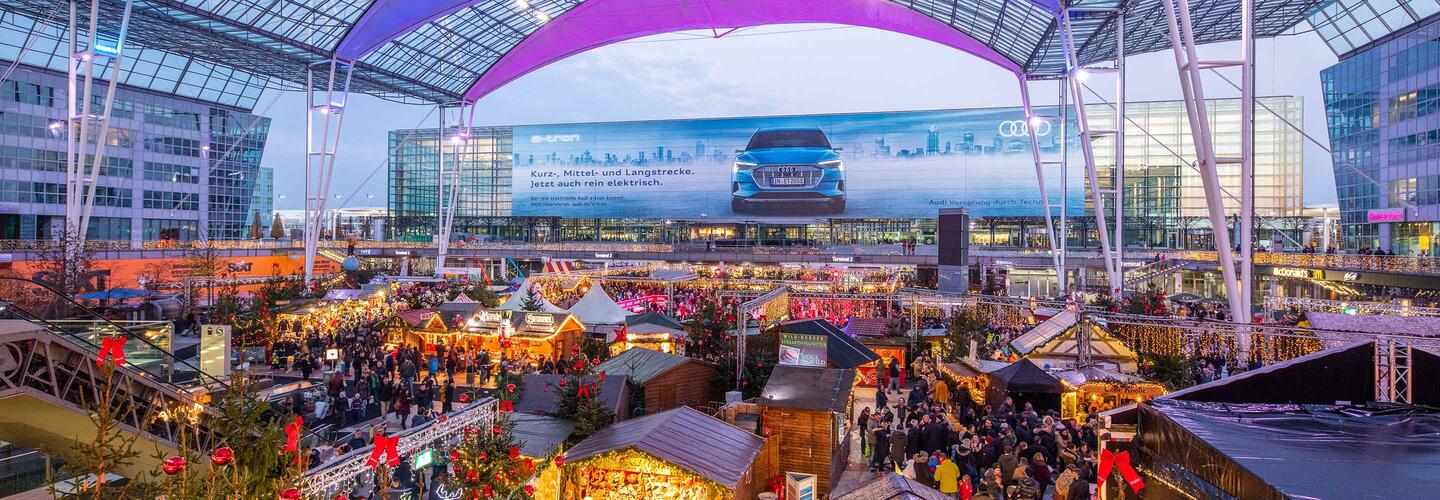 Wintermarkt am Flughafen Großer Winter- und Weihnachtsmarkt im München Airport Center mit bunten Lichtern, festlichen Ständen und vielen Besucherinnen und Besuchern unter dem markanten Glasdach. Die lebendige Atmosphäre, warmes Licht und die marktähnliche Kulisse vermitteln echte Winterzauber-Stimmung am Flughafen München.