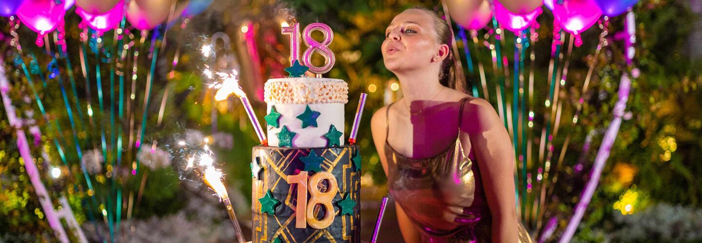 Geburtstagsparty Eine junge Frau steht lächelnd vor einer bunt dekorierten Geburtstagstorte mit der Zahl 18, umgeben von glitzernden Lichtern und farbenfrohen Partydekorationen. Die Szene wirkt festlich, energiegeladen und voller Vorfreude.