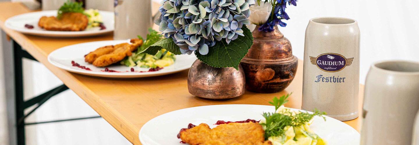 Bayerisches Fest Ein Tisch mit traditionell bayerischen Gerichten wie panierten Schnitzeln und frischen Salaten, daneben ein Steinkrug mit ‚Flughafen München‘-Aufdruck sowie eine Vase mit Blumen. Die Szene wirkt gemütlich, einladend und typisch bayerisch.