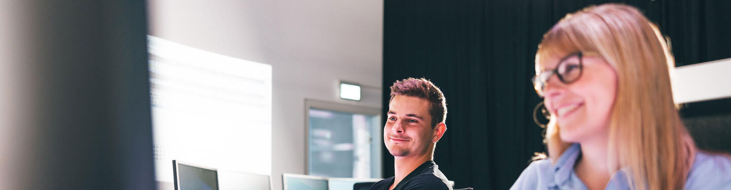 Duales Studium Informatik am Flughafen München Ein junger Mann und eine junge Frau sitzen lächelnd an Computerarbeitsplätzen und arbeiten gemeinsam an IT-Projekten am Flughafen München; ruhige, positive Lern- und Arbeitsatmosphäre.