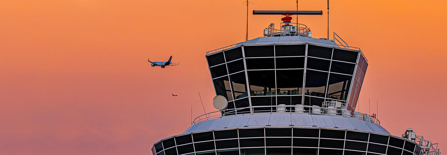 Unsere Marke: Wofür wir stehen Flugzeug im Landeanflug vor einem intensiv orangenen Abendhimmel. Im Vordergrund die Silhouette des Münchner Flughafentowers. Die Szene wirkt beeindruckend, ruhig und gleichzeitig dynamisch durch das herannahende Flugzeug.