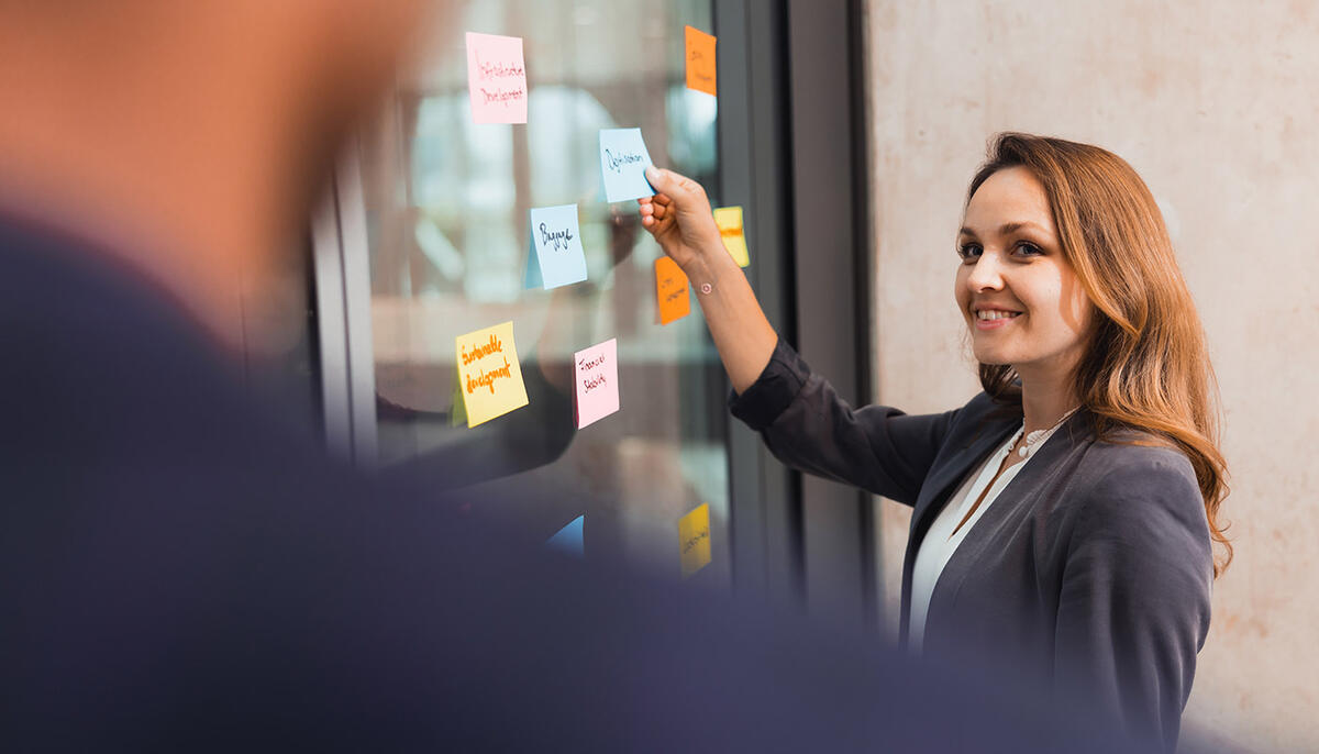 Workshop AirportAcademy Frau klebt Postis an Glasscheibe und blickt zu ihrem Kollegen.