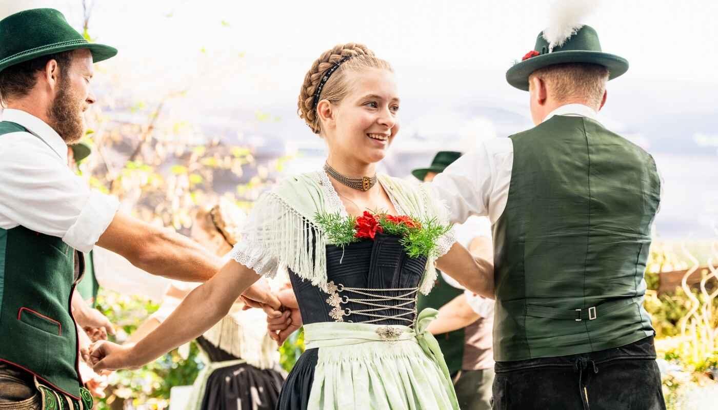 Bayerische Tracht und Volkstanz Eine junge Frau im Dirndl lächelt strahlend, während sie mit anderen in Tracht im Kreis tanzt, Hände verbunden, lebendige Tradition und Lebensfreude in der warmen Sonne.