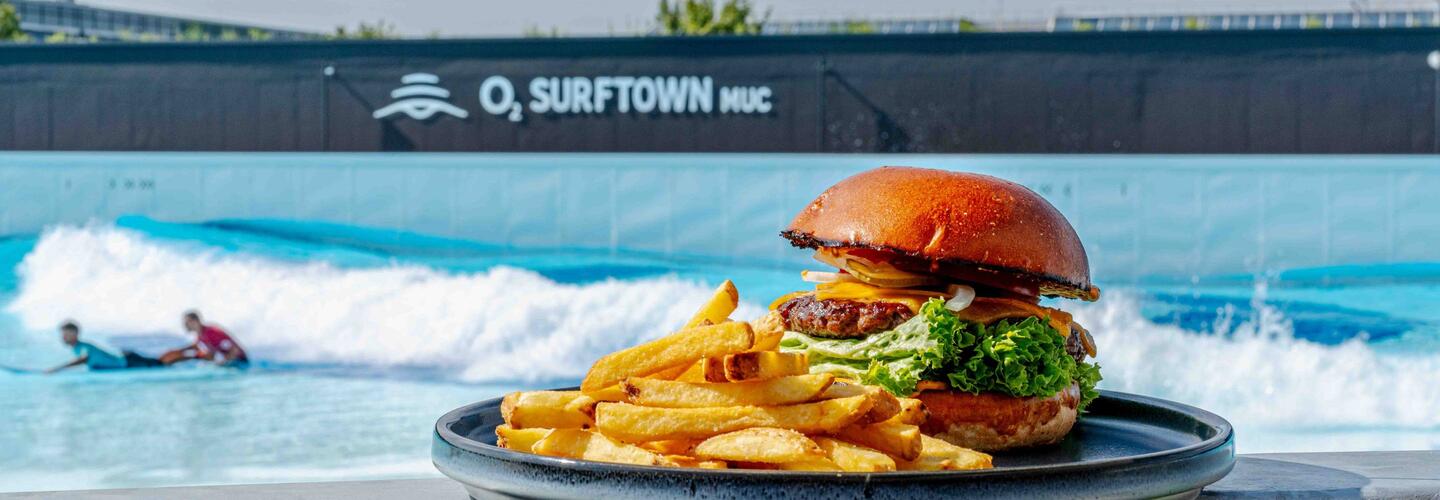 Surftown Lookout - Burger with surf wave in the background Burger and fries on a plate in front of the O₂ Surftown MUC surfing facility, with surfers riding the artificial wave in the background.