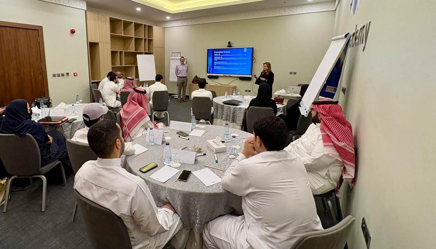 PXM Training Session in Riyadh Präsentation mit Teilnehmenden in traditioneller arabischer Kleidung in einem Konferenzraum am King Khalid International Airport.