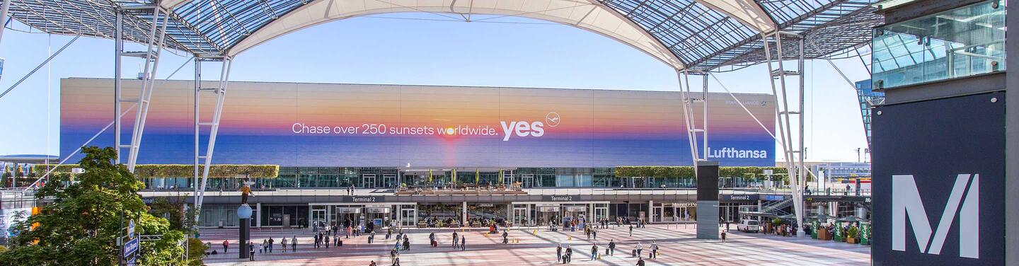 Lufthansa Westfassade - Sonnenuntergang Die Westfassade des Terminals 2 am Flughafen München ist mit einem Motiv der Lufthansa bespielt. Darauf ist ein Sonnenaufgang zu sehen sowie der Schriftzug "Chase over 250 sunsets worldwide - yes".