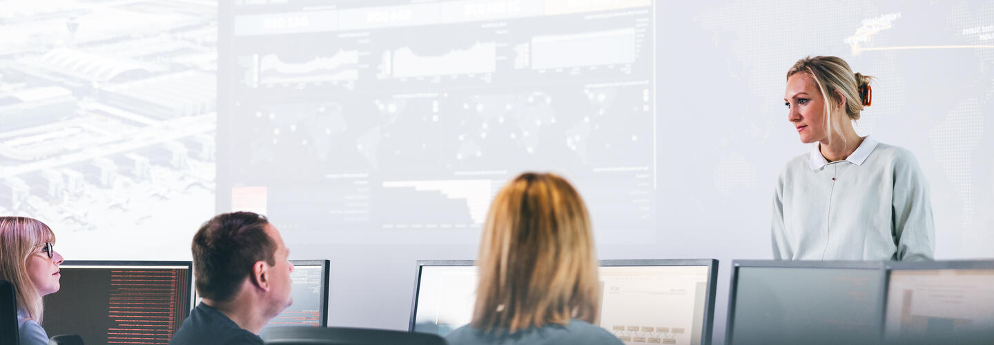 CSR Schulung im ISH Trainerin steht vor Monitorwänden und spricht zu Schulungsteilnehmern, die vor ihr an Bildschirmen sitzen.