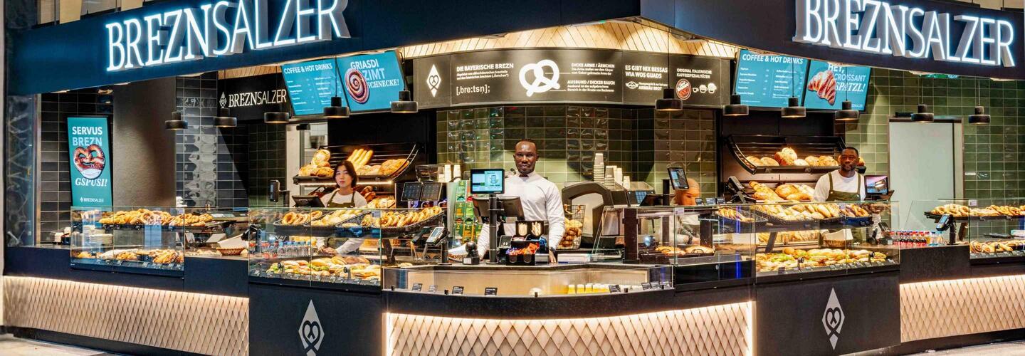 Keyvisual Allresto Breznsalzer Modern gestalteter Breznsalzer-Verkaufsstand mit beleuchtetem Tresen und großer Auswahl an Brezeln und Backwaren; drei Mitarbeitende stehen hinter der Theke und bedienen Kunden.