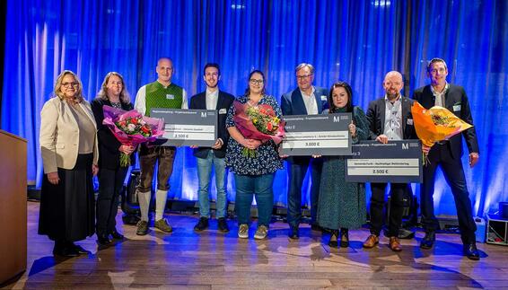 Gruppenfoto beim Regionalempfang 2025: Mehrere Personen stehen auf einer Bühne vor blau beleuchtetem Hintergrund, halten große symbolische Spendenschecks und Blumensträuße und werden für ihr ehrenamtliches Engagement in der Region von der Flughafen München GmbH ausgezeichnet.