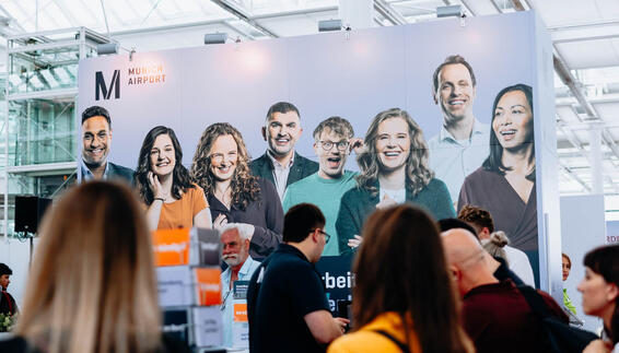Das Bild zeigt den Messestand des Flughafens München auf der „jobmesse munich airport“ mit einem großen Banner, auf dem mehrere Mitarbeitende sowie das Logo „Munich Airport“ abgebildet sind. Im Vordergrund sind Messebesucher:innen zu sehen.