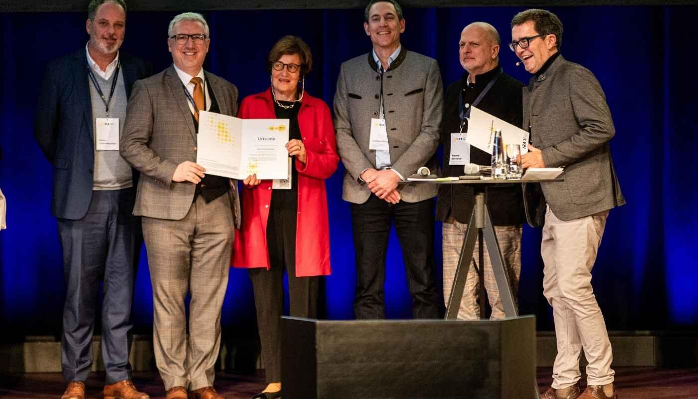 Auszeichnung der Nachbarschaftsregion echs Personen stehen auf einer Bühne vor blau beleuchtetem Hintergrund. Zwei von ihnen halten eine Urkunde, die gemeinsam präsentiert wird. Die Gruppe wirkt feierlich und lächelt, während rechts ein Stehtisch mit Mikrofonen und Unterlagen steht.