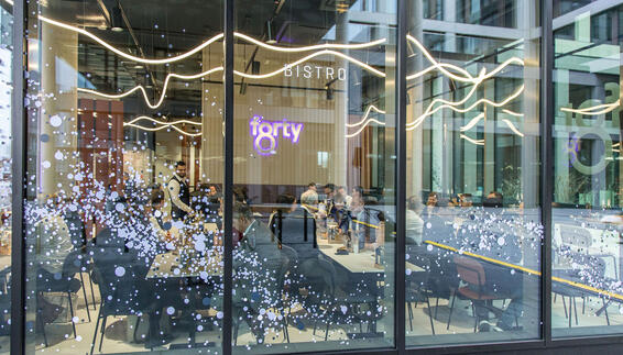 Blick von Außen durch die Fensterscheibe des Bistro forty8 im LabCampus. Man sieht ein modern eingerichtetes Bistro mit abendlicher Beleuchtung. Die Stimmung ist entspannt. 