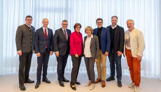 37. Plenumssitzung des Nachbarschaftsbeirats Gruppenfoto von acht Erwachsenen, die in einer Reihe vor einem hellen, lichtdurchlässigen Vorhang stehen. Die Personen tragen überwiegend formelle Kleidung, einige Anzüge, Blazer oder traditionelle Jacken. Zwei Frauen stehen in der Mitte, flankiert von sechs Männern. Alle blicken lächelnd in die Kamera.