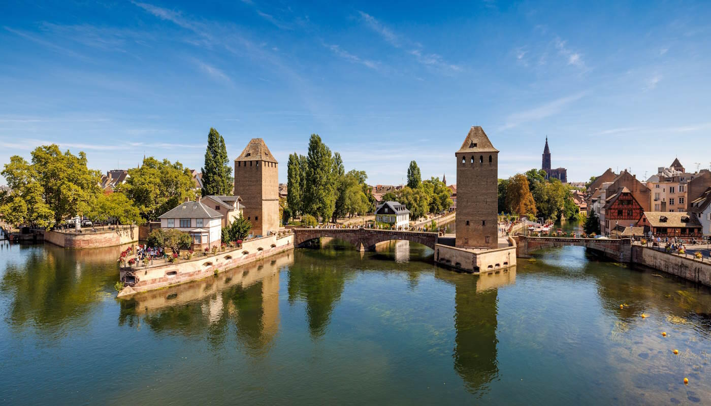 Ponts Couverts Brücke in Straßburg, Frankreich Idyllische Ansicht der Ponts Couverts Brücke in Straßburg, Frankreich mit dem ruhigen Fluss Ill. Im Hintergrund sind die massiven, mittelalterlichen Wehrtürme, die sich im Wasser spiegeln. Entlang der Ufer wachsen Bäume, dahinter sind traditionelle Häuser zu sehen.