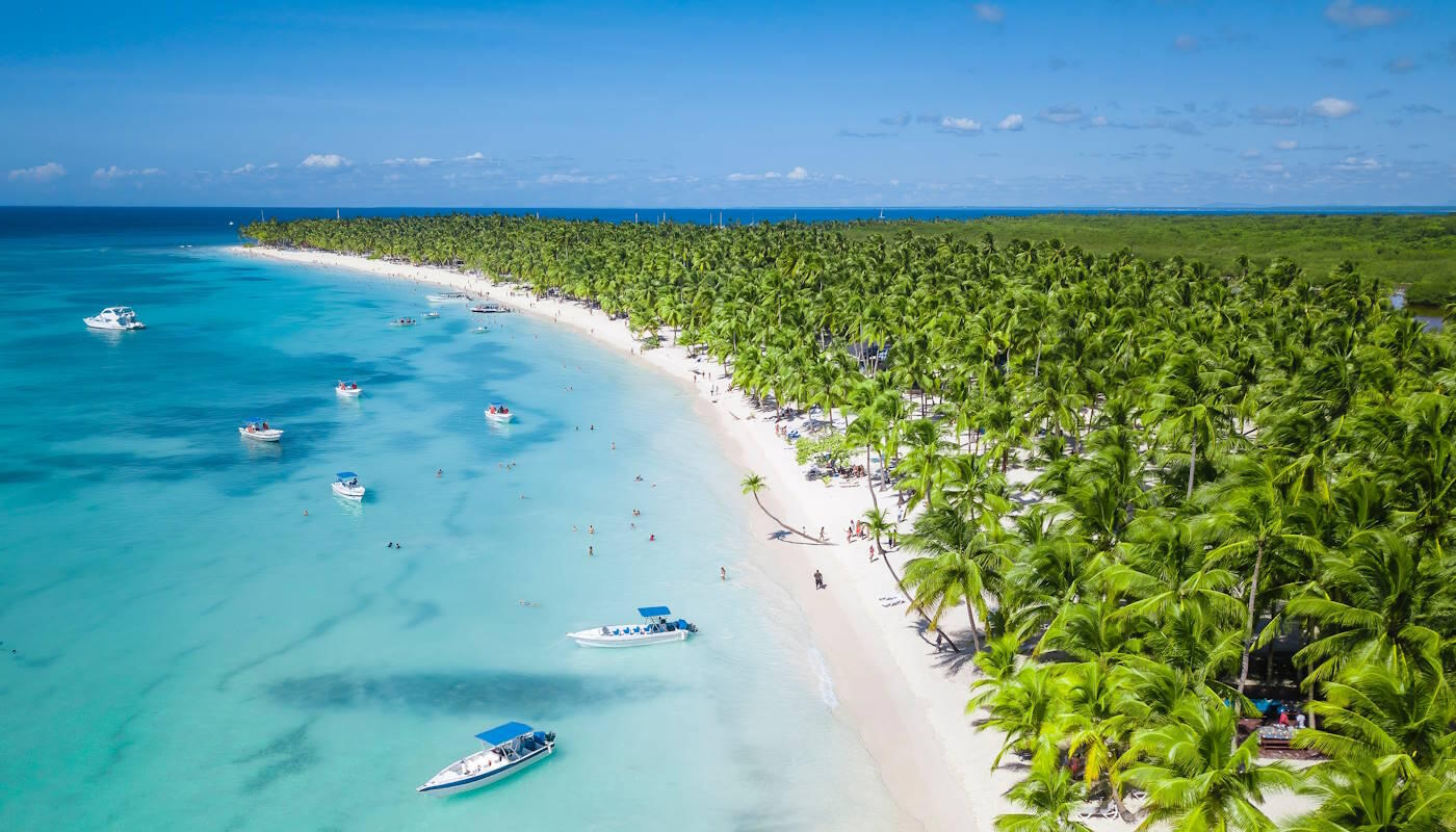 Soana Island in der Dominikanischen Republik Soana Island in der Dominikanischen Republik. Ein langer, geschwungener Sandstrand mit feinem, hellem Sand zieht sich entlang einer Küste. Direkt hinter dem Strand steht ein dichter Palmenwald.Das Wasser ist klar und türkisfarben. Im flachen Wasser und etwas weiter draußen liegen mehrere kleine Boote und Motorboote.