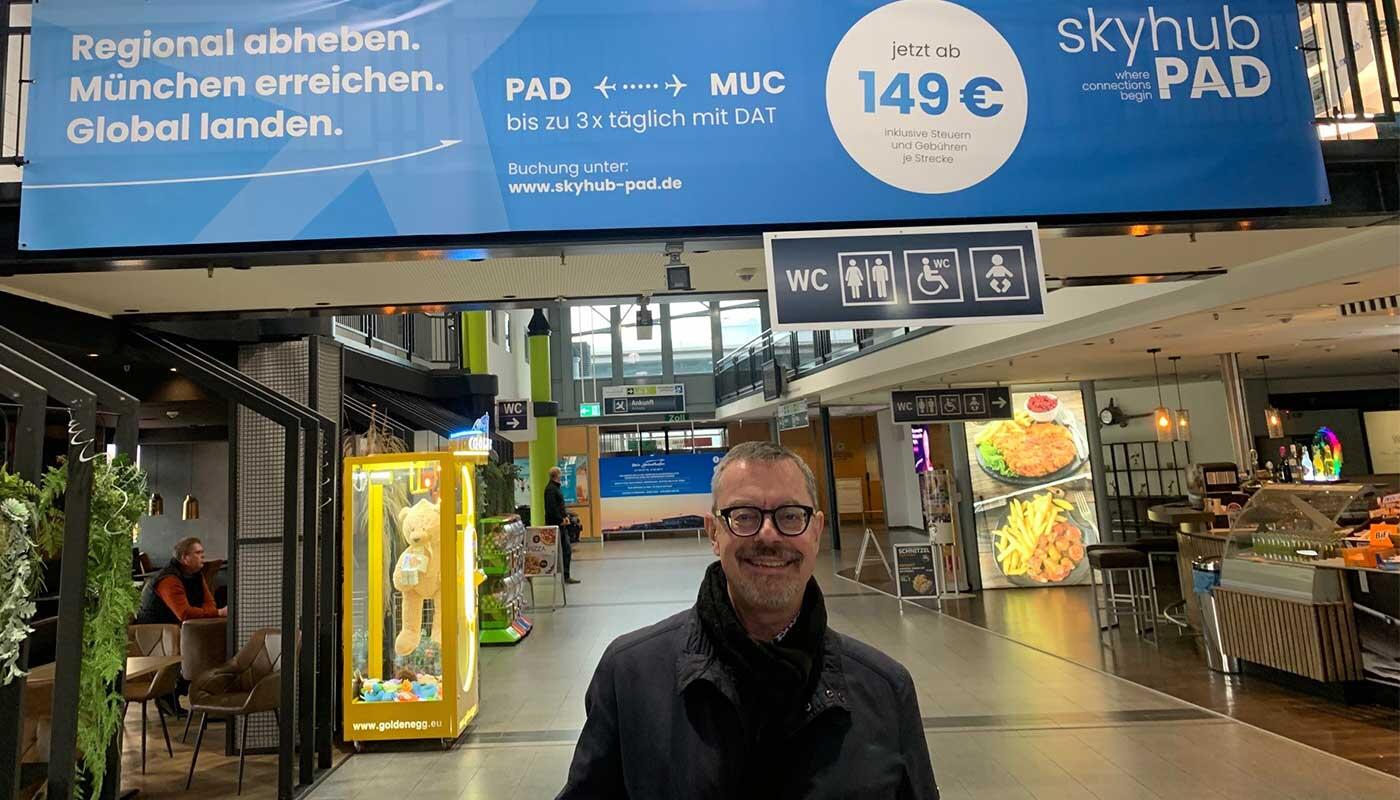 Paderborn Insider Stefan Metzler Stefan Metzler im Innenbereich des Flughafens Paderborn/Lippstadt. Er steht entspannt im Terminal, im Hintergrund sind Gastronomie, Sitzbereiche und Hinweisschilder zu sehen.