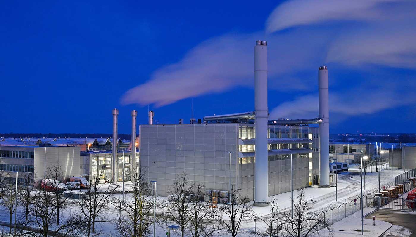 Wärmeversorgung – mit Volldampf gegen die Kälte Heiz- und Energiezentrale am Flughafen München bei Nacht, umgeben von Schnee, mit beleuchteten Gebäuden und aufsteigendem Dampf aus Schornsteinen.“