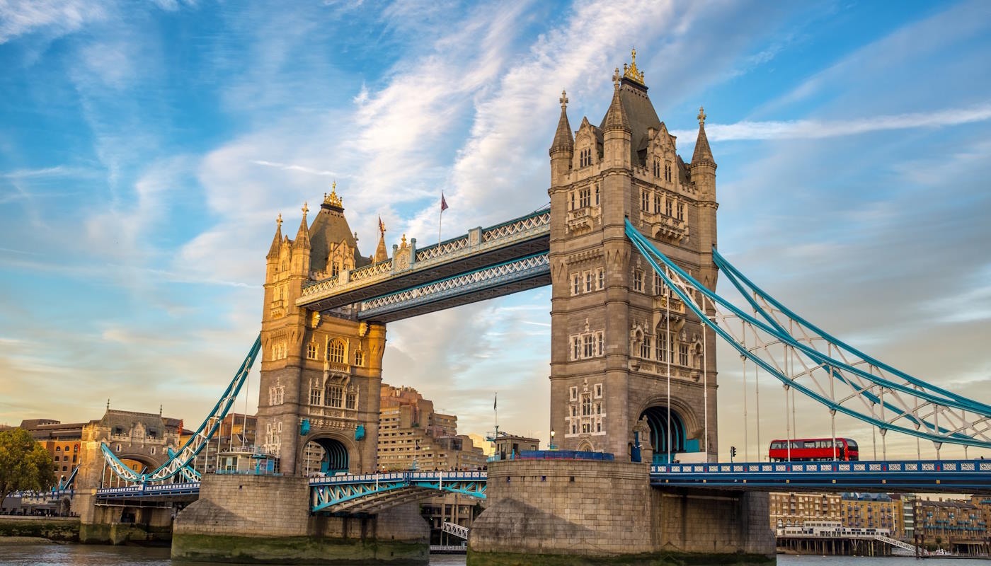 Tower Bridge, London, UK Die markante Tower Bridge in London, eine Klappbrücke, überspannt die Themse und besteht aus zwei massiven, steinernen Türmen im neugotischen Stil, die durch einen oberen Fußgängersteg miteinander verbunden sind. Die Brückenkonstruktion kombiniert grauen Stein mit blau lackierten Stahlträgern. Im Vordergrund fließt ruhig der Fluss, während im Hintergrund städtische Gebäude zu erkennen sind. Der Himmel ist weitgehend klar. Auf der Fahrbahn ist ein roter Doppeldeckerbus zu sehen.