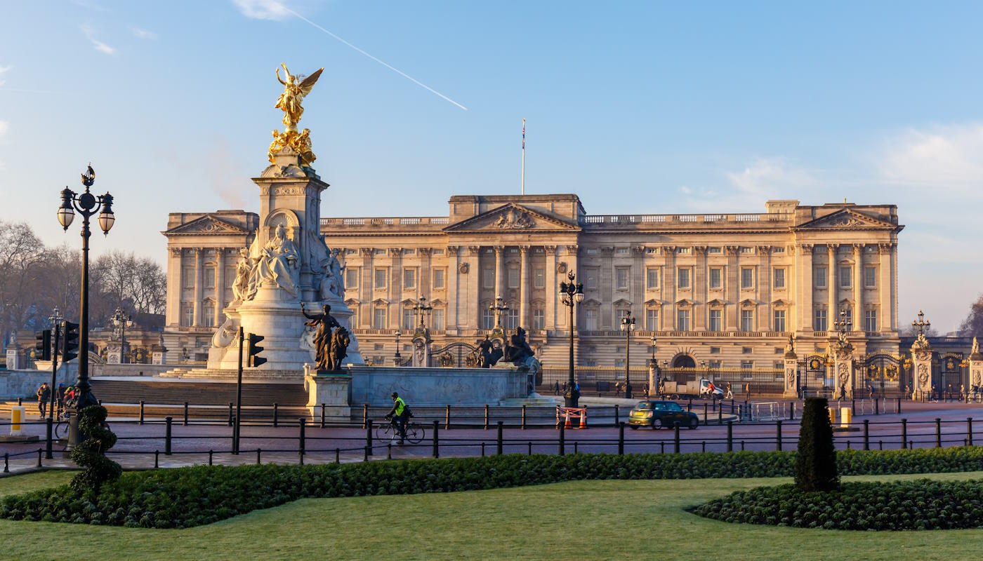 Buckingham Palace, London Buckingham Palace in London bei warmem, vermutlich spätnachmittäglichem Licht. Im Vordergrund steht das Victoria Memorial – ein großes weißes Denkmal mit goldener Statue an der Spitze. Die vergoldete Figur oben glänzt im Sonnenlicht. Vor dem Denkmal verläuft eine Straße, auf der ein Fahrradfahrer unterwegs ist; außerdem sind einige Autos zu sehen. Schwarze Laternen und Absperrungen säumen den Platz. Im unteren Bildbereich erkennt man gepflegte Grünflächen und symmetrisch angelegte Beete. Der Himmel ist klar und blau.