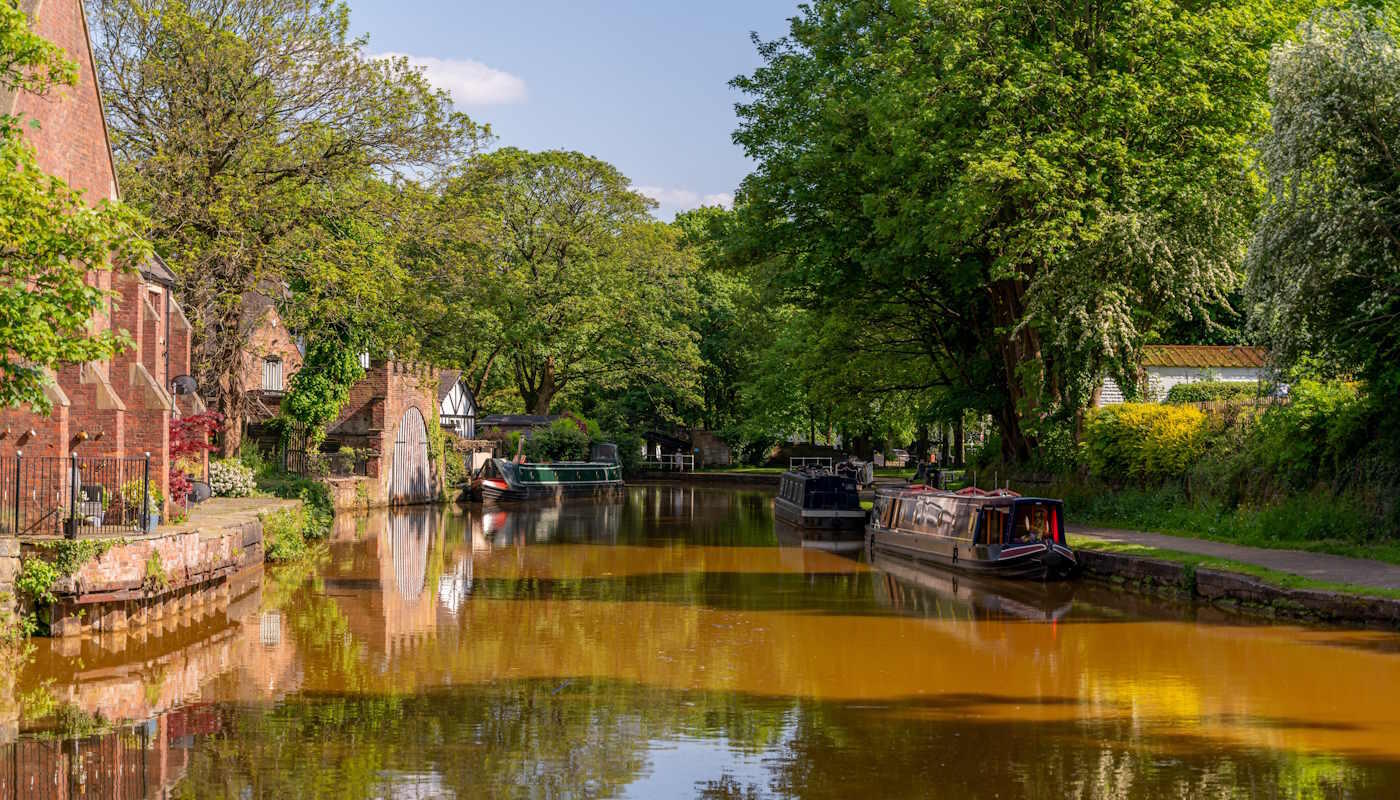Boote auf dem Bridgwater-Kanal in Worsley, Manchester Boote auf dem Bridgwater-Kanal in Worsley, Manchester. Ein ruhiger Kanal mit mehreren langen, schmalen Booten, die am Ufer festgemacht sind. Üppige grüne Bäume spiegeln sich im Wasser, ein kleines Backsteinhaus und eine steinerne Brücke rahmen die friedliche Umgebung ein.