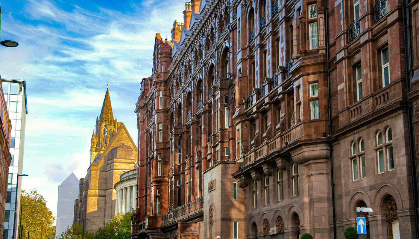 Peter Street, Manchester, UK Großes, reich verziertes Gebäude in der Peter Street in Manchester aus rötlich-braunem Stein mit vielen Fenstern, Ornamenten und Bögen an der Fassade. Im Hintergrund steht eine imposante Kirche mit einem hohen, spitzen Turm. Der Himmel ist klar.