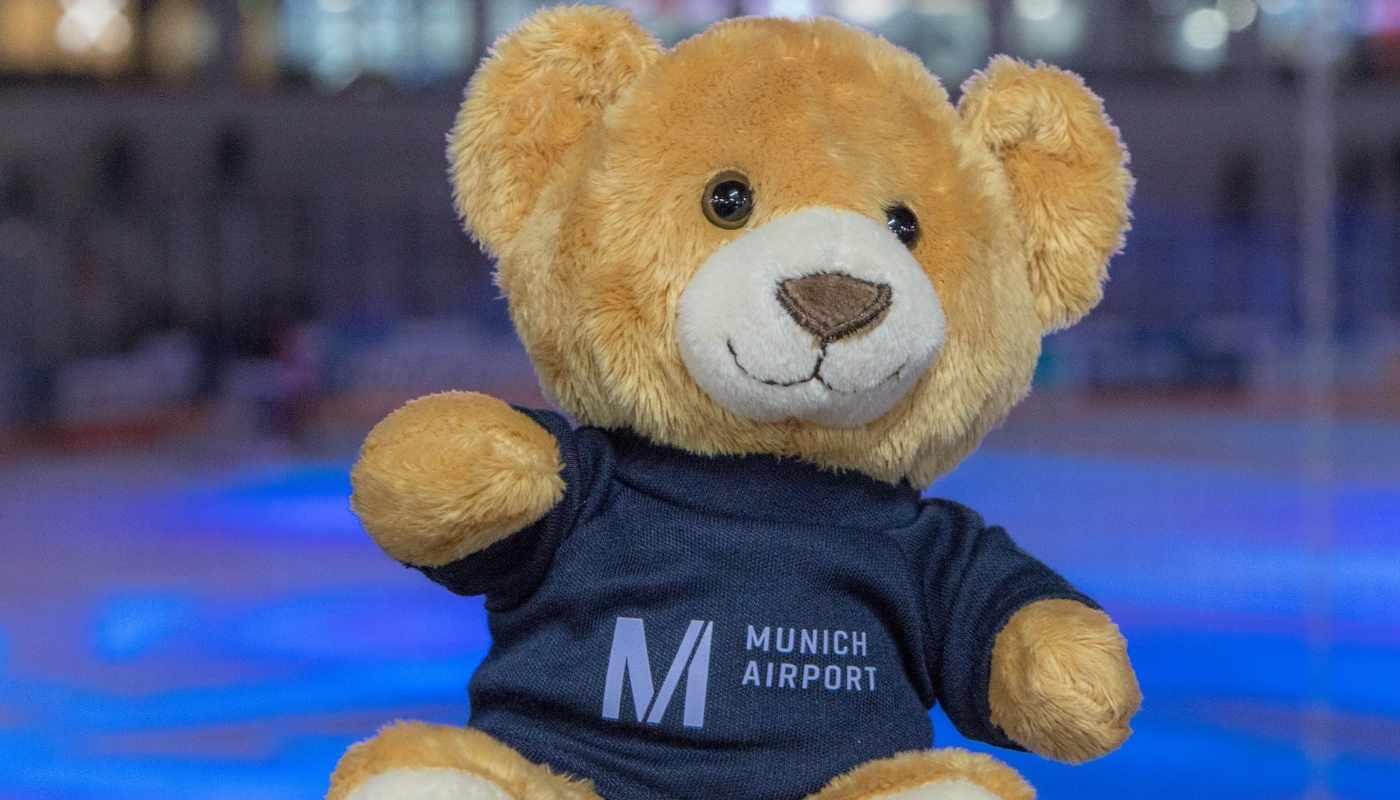 Teddybär mit Munich Airport Shirt beim Benefiz-Eishockey – Teddy Bear Toss Aktion Teddybär mit dunkelblauem Munich-Airport-Shirt vor unscharfem Hintergrund einer Eishockeyarena während einer Benefizaktion.