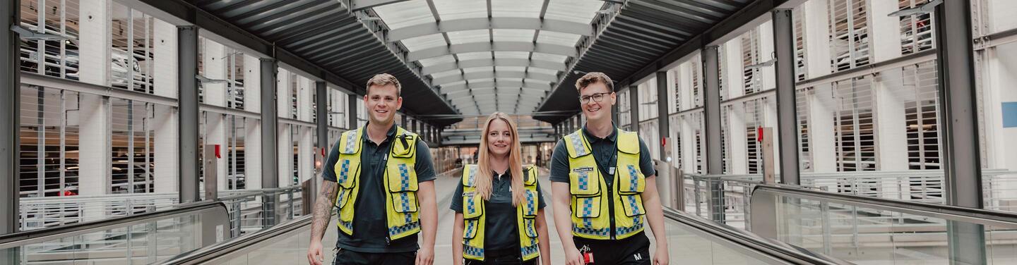 Ausbildung Fachkraft für Schutz und Sicherheit Drei Personen in gelben Sicherheitswesten gehen durch einen überdachten, modernen Übergang mit Glas und Metallstrukturen.