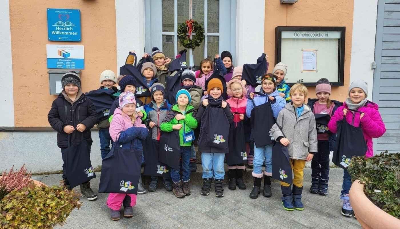 Flughafen München spendet Büchertaschen an Grundschule Velden Erstklässler der Grundschule Velden stehen vor dem Schulgebäude und halten vom Flughafen München gespendete dunkle Büchertaschen in die Kamera; die Kinder tragen Winterkleidung und lächeln bei einer offiziellen Übergabeaktion im Außenbereich der Schule.