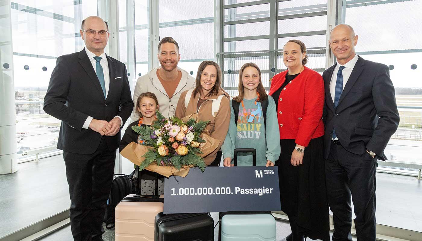Flughafen München begrüßt 1.000.000.000. Passagier Gruppe mit Koffergepäck am Flughafen hält ein Schild für den milliardsten Passagier und einen Blumenstrauß