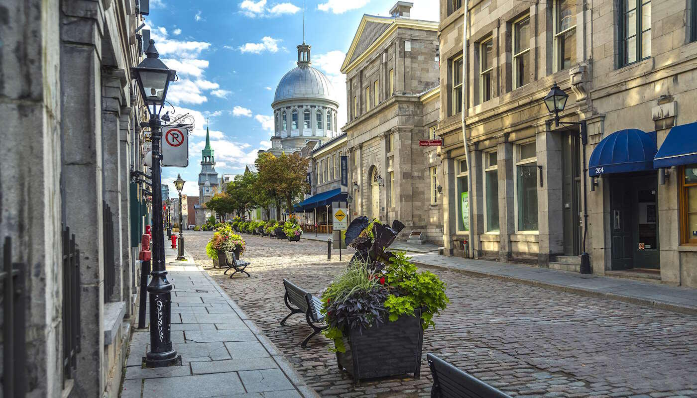 Altstadt Montreal, Kanada Kopfsteinpflasterstraße mit historischen Gebäuden, Laternen und Blumenarrangements in der Altstadt von Montreal, Kanada. Im Hintergrund ist die markante Kuppel eines Gebäudes zu sehen, typisch für Old Montreal passt. Die Straße ist sauber und gepflegt, der Himmel ist blau.