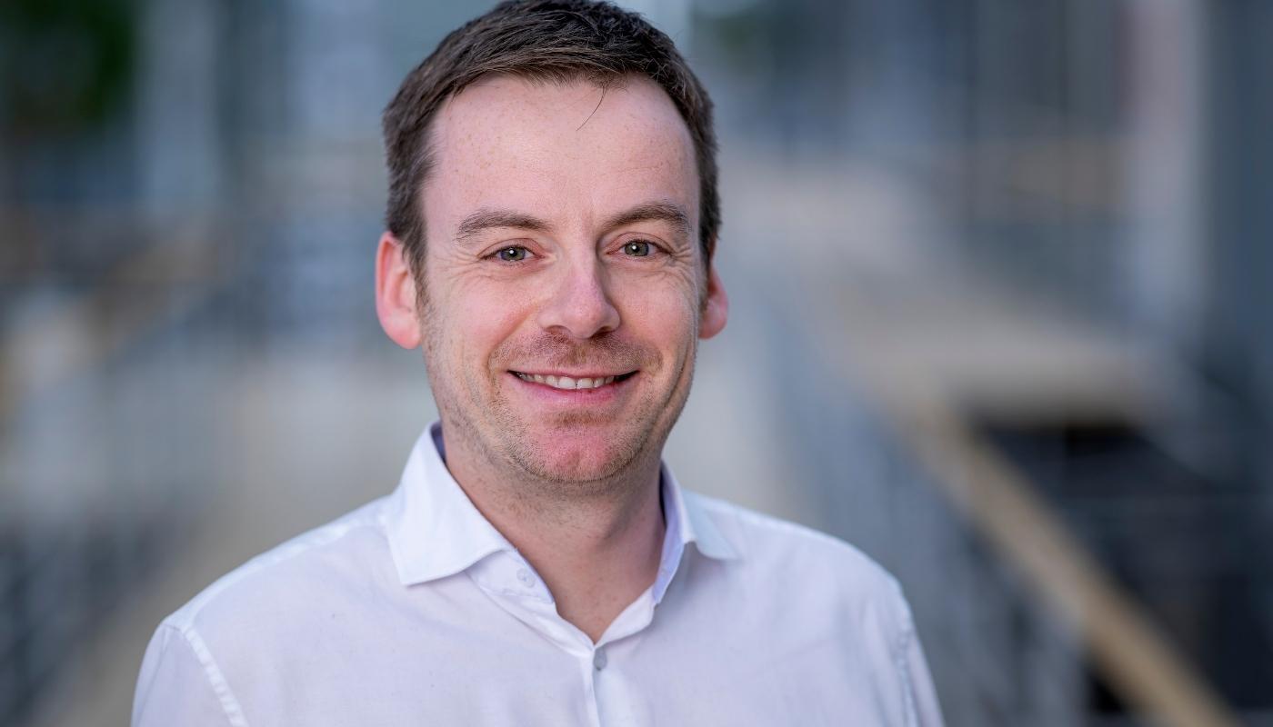 Interview mit Sebastian Ibel Porträt eines lächelnden Mannes in weißem Hemd vor unscharfem Hintergrund im urbanen Umfeld. Der Mann blickt direkt in die Kamera und wirkt freundlich und professionell.