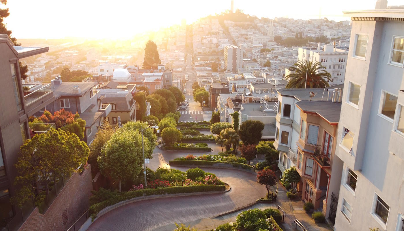 Lombard Street in San Francisco, USA Lombard Street in San Francisco, USA. Eine steile Straße mit mehreren engen Kurven, die von üppigen Pflanzen und gepflegten Gärten und Häusern gesäumt wird. Dahinter breitet sich eine sonnig beleuchtete Skyline aus, die sich bis zum Horizont erstreckt.