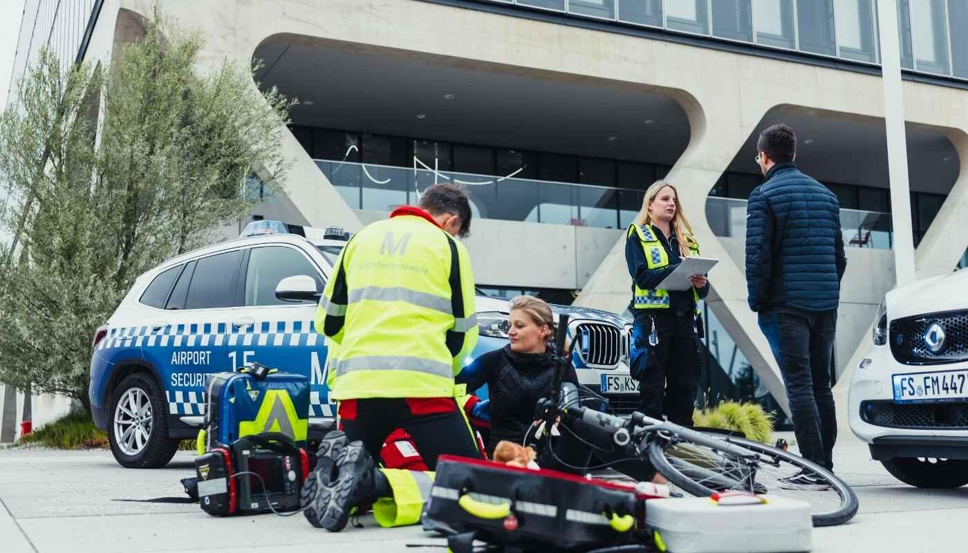 Notfallambulanz Flughafen München – Einsatz vor Ort Rettungskräfte der Notfallambulanz am Flughafen München versorgen eine verletzte Person neben einem Einsatzfahrzeug. Medizinische Ausrüstung liegt bereit, während das Team professionell vor dem Terminal arbeitet.
