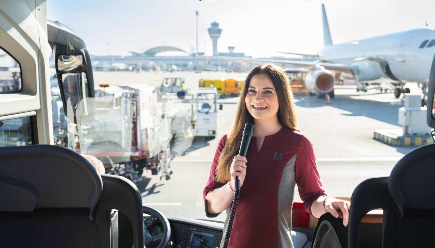 Flughafen-Erlebnis für Familien – Einblicke direkt auf dem Vorfeld Eine Mitarbeiterin steht im Bus auf dem Vorfeld eines Flughafens und spricht in ein Mikrofon, während im Hintergrund Flugzeuge und Servicefahrzeuge zu sehen sind.