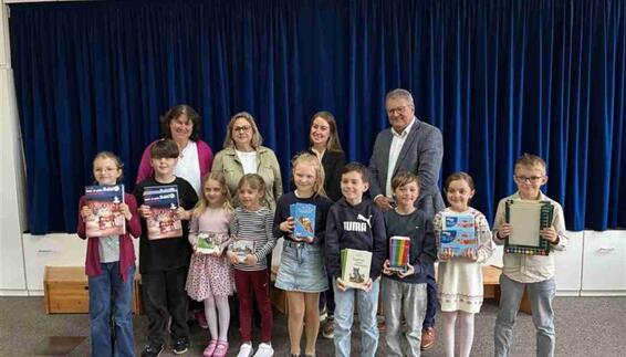 Grundschulkinder halten ihre neuen Bücher in den Händen und blicken mit vier Erwachsenen gemeinsam vor einer Bühne lächelnd in die Kamera. Im Hintergrund ein dunkelblauer, geschlossener Bühnenvorhang