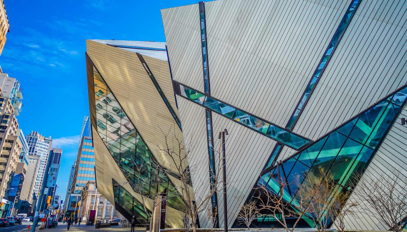 Royal Ontario Museum in Toronto, Kanada Das Bild zeigt den markanten modernen Anbau „Michael Lee‑Chin Crystal“ des Royal Ontario Museum in Toronto, Kanada. Zu sehen ist die kantige Glas‑ und Aluminiumfassade an einer belebten Innenstadtstraße mit umliegenden Hochhäusern. Der Himmel ist klar und blau.