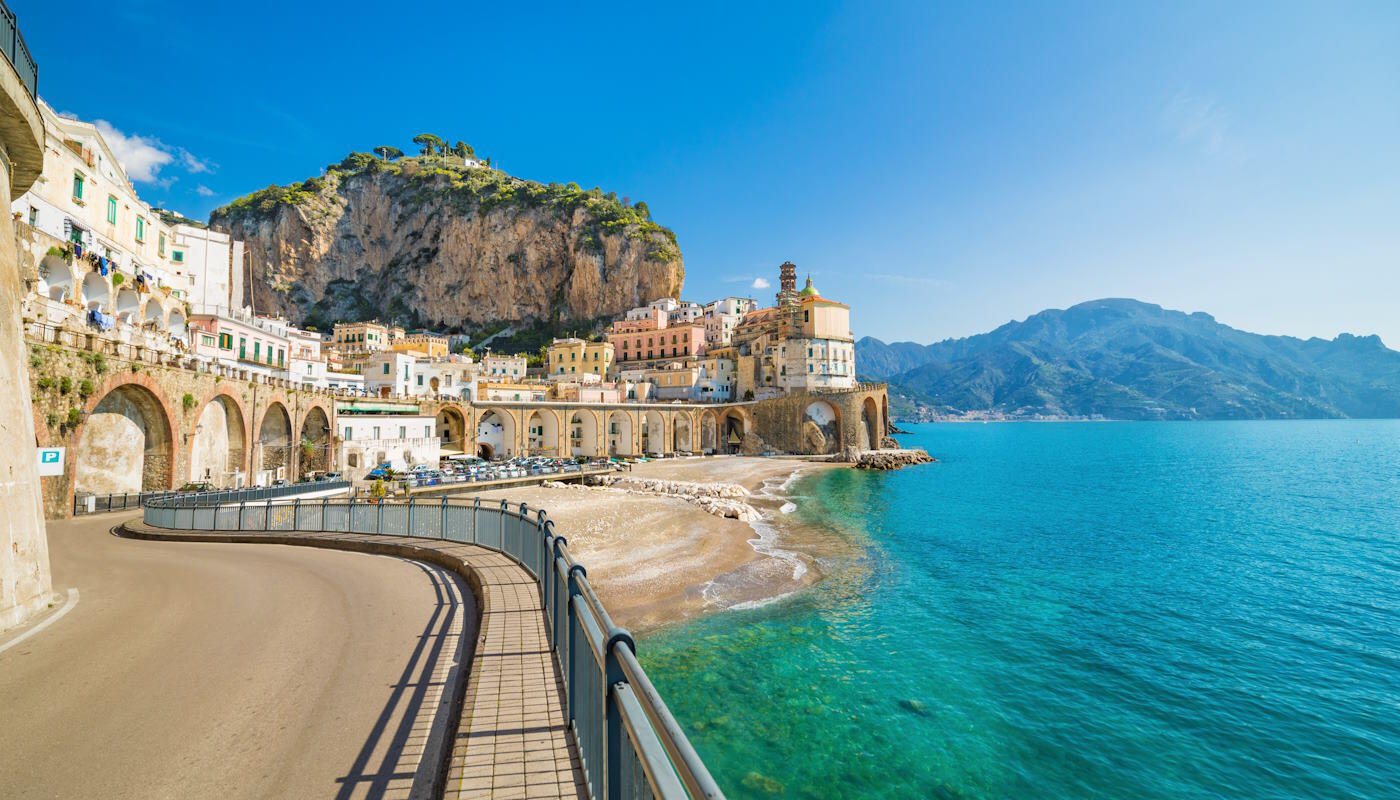 Amalfiküste, Italien Die Küstenstadt Atrani an der Amalfiküste in Italien mit pastellfarbenen Häusern, die direkt an steilen Felsen über dem türkisblauen Meer gebaut sind. Die geschwungene Straße und die Arkaden entlang der Küste sind typisch mediterran, mit sonnigem Himmel und dramatischer Bergkulisse im Hintergrund.