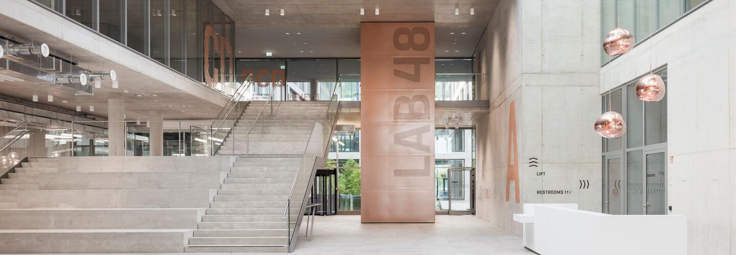 Moderne Architektur und inspirierende Business-Atmosphäre im LabCampus München Moderner, lichtdurchfluteter Eingangsbereich des LabCampus am Flughafen München mit großer Treppe, hohen Glasfronten, stilvollen Kupfer-Designelementen und offener Business-Atmosphäre.