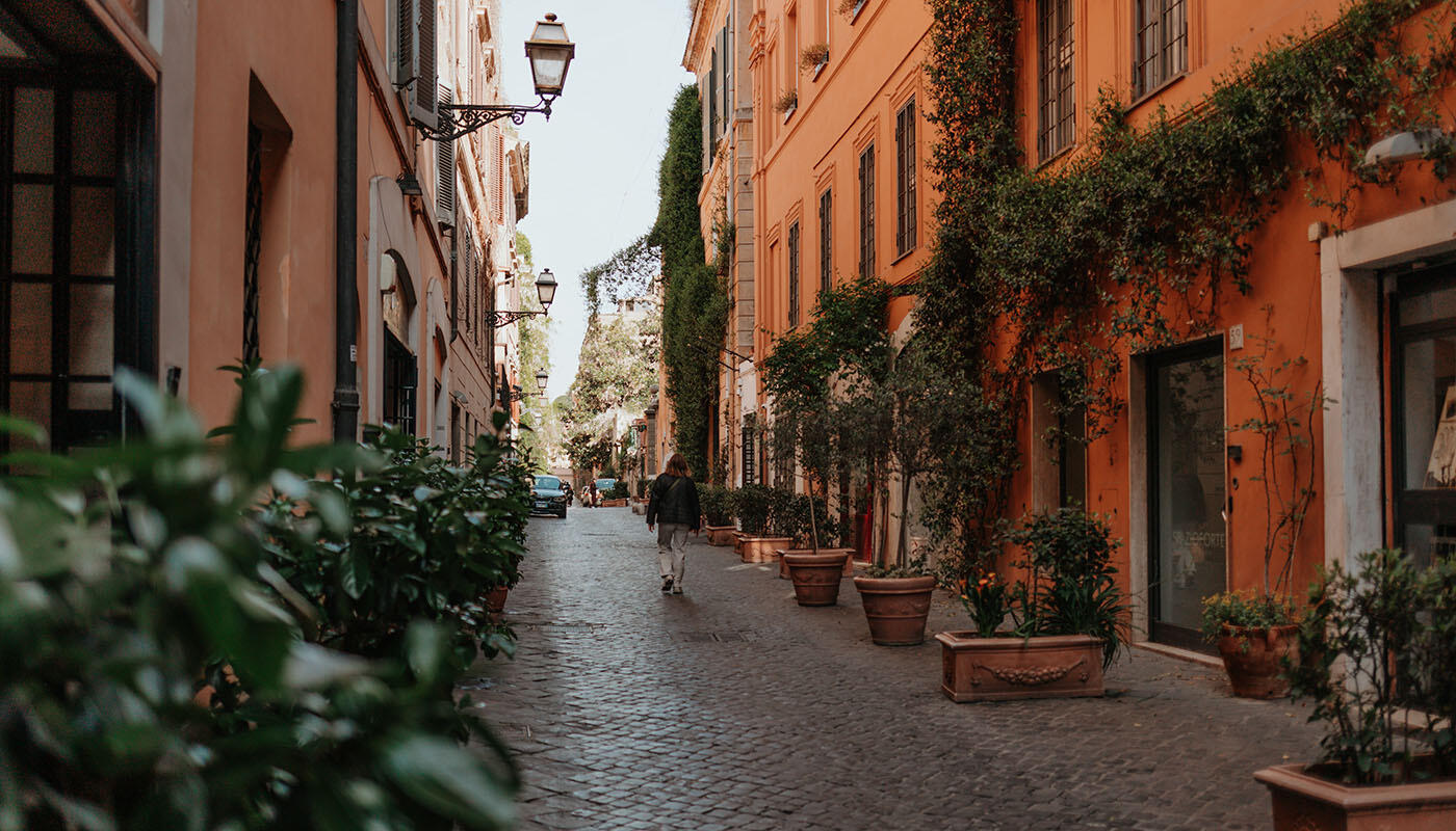 Via Margutta Gasse Rom Ruhige, enge Gasse mit Kopfsteinpflaster und warmfarbenen Häuserfassaden. Viele Pflanzen und Blumentöpfe säumen den Weg. Eine Person geht in der Mitte der Straße entlang.