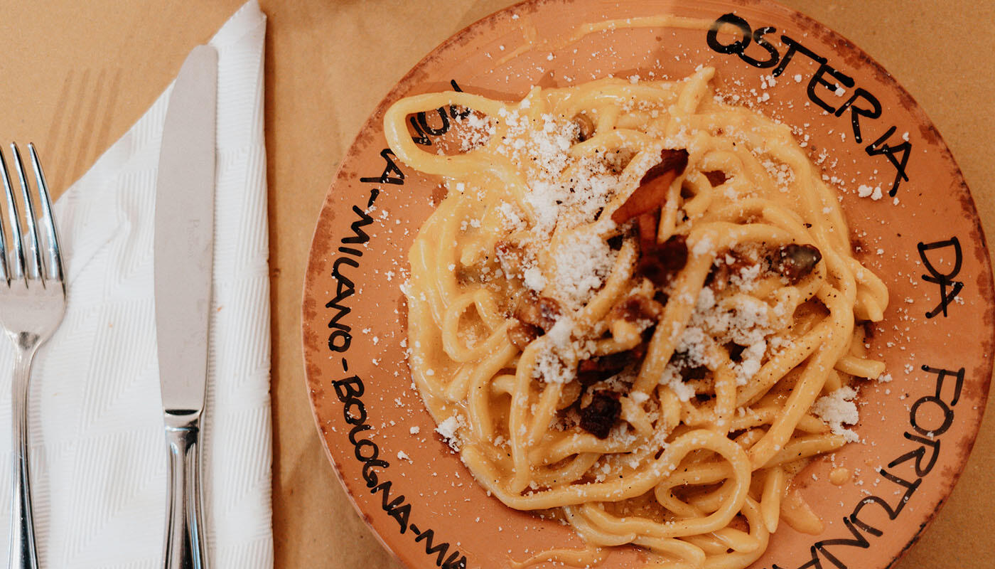 Osteria da fortunata Carbonara Teller mit cremiger Pasta, bestreut mit geriebenem Käse und Speckstücken. Daneben liegen Besteck und eine Serviette auf dem Tisch. Der Teller trägt die Aufschrift „Osteria da Fortunata“.