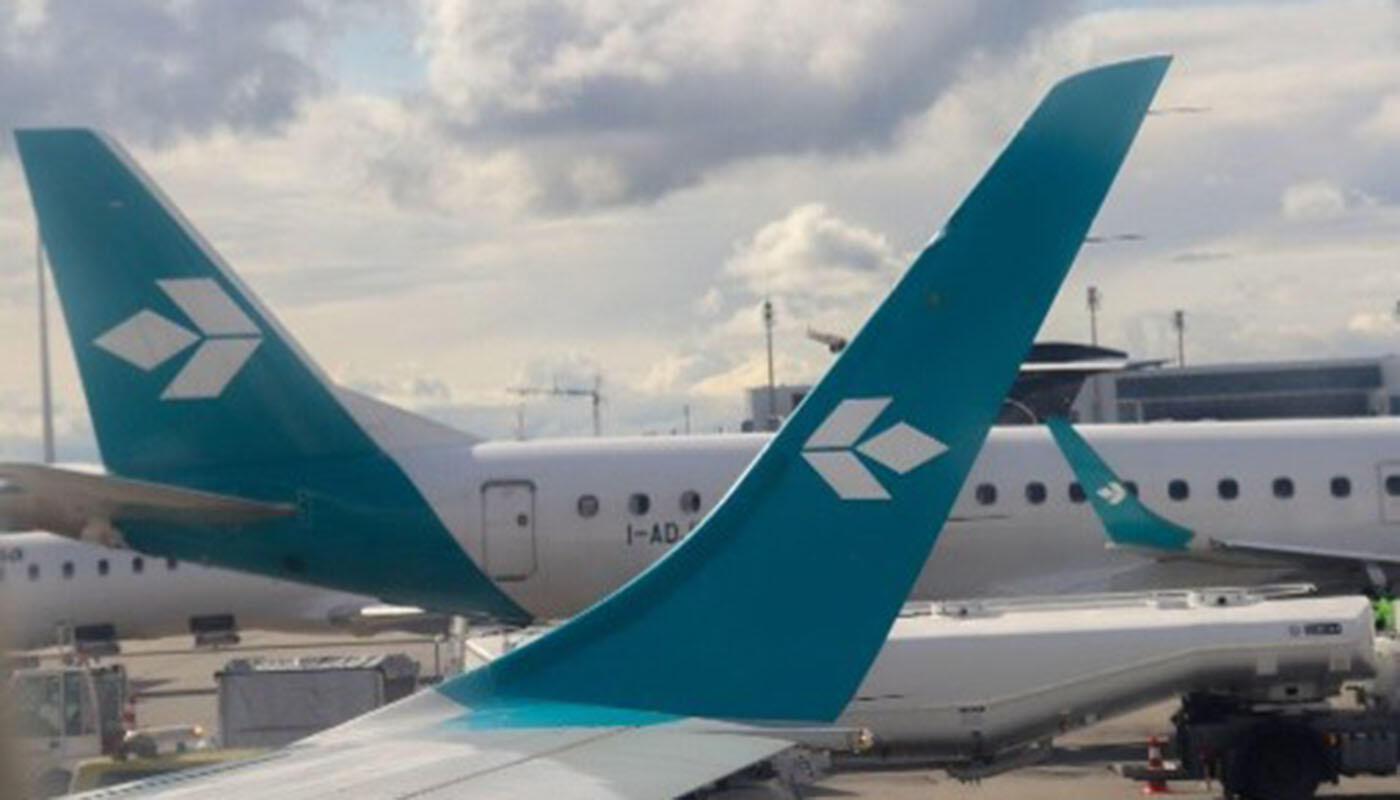 Air Dolomiti Mehrere Flugzeuge stehen auf dem Rollfeld des Flughafens. Die Leitwerke sind in Türkis gehalten und tragen ein geometrisches Logo der Airline Air Dolomiti. Im Hintergrund sind Flughafenanlagen und Fahrzeuge sichtbar.