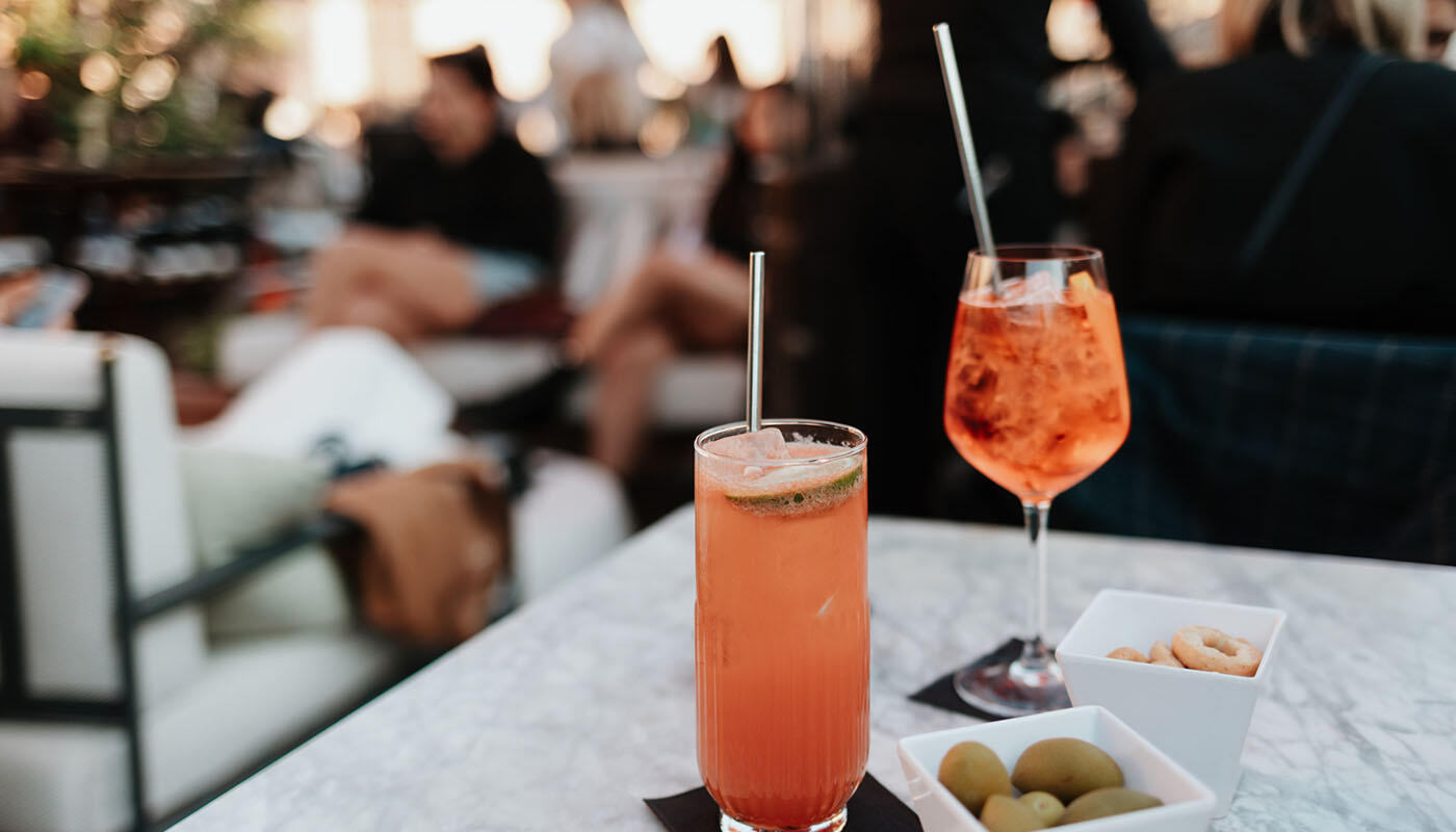 Drinks in Rom Zwei farbige Cocktails stehen auf einem Marmortisch in einer Bar. Dazu werden kleine Snacks wie Oliven und Gebäck in Schalen serviert. Im Hintergrund sitzen Gäste in einer entspannten Atmosphäre.