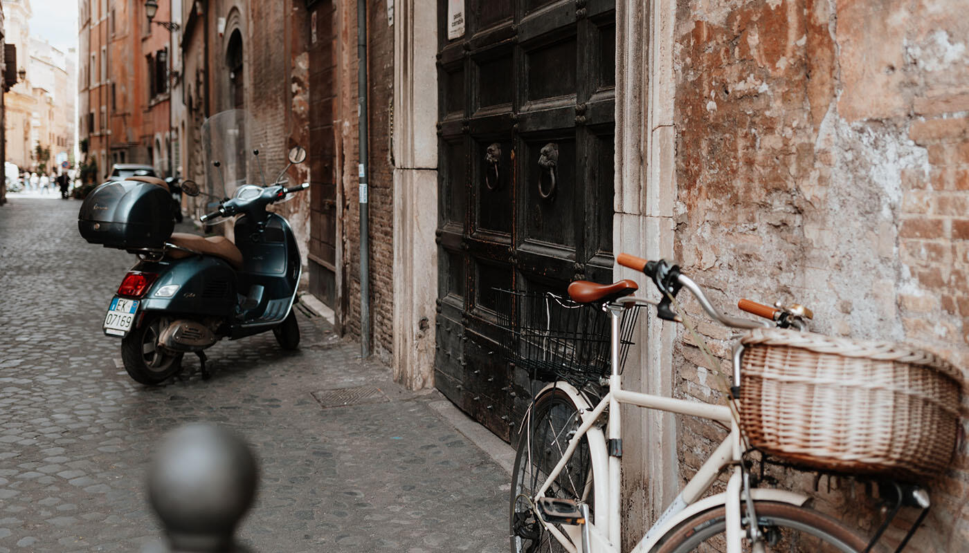 Rom Mood Ein Fahrrad mit Korb lehnt an einer alten Steinwand in einer schmalen Gasse. Daneben steht ein Motorroller auf dem Kopfsteinpflaster. Die Umgebung zeigt typische, leicht verwitterte Gebäude im Stadtbild.